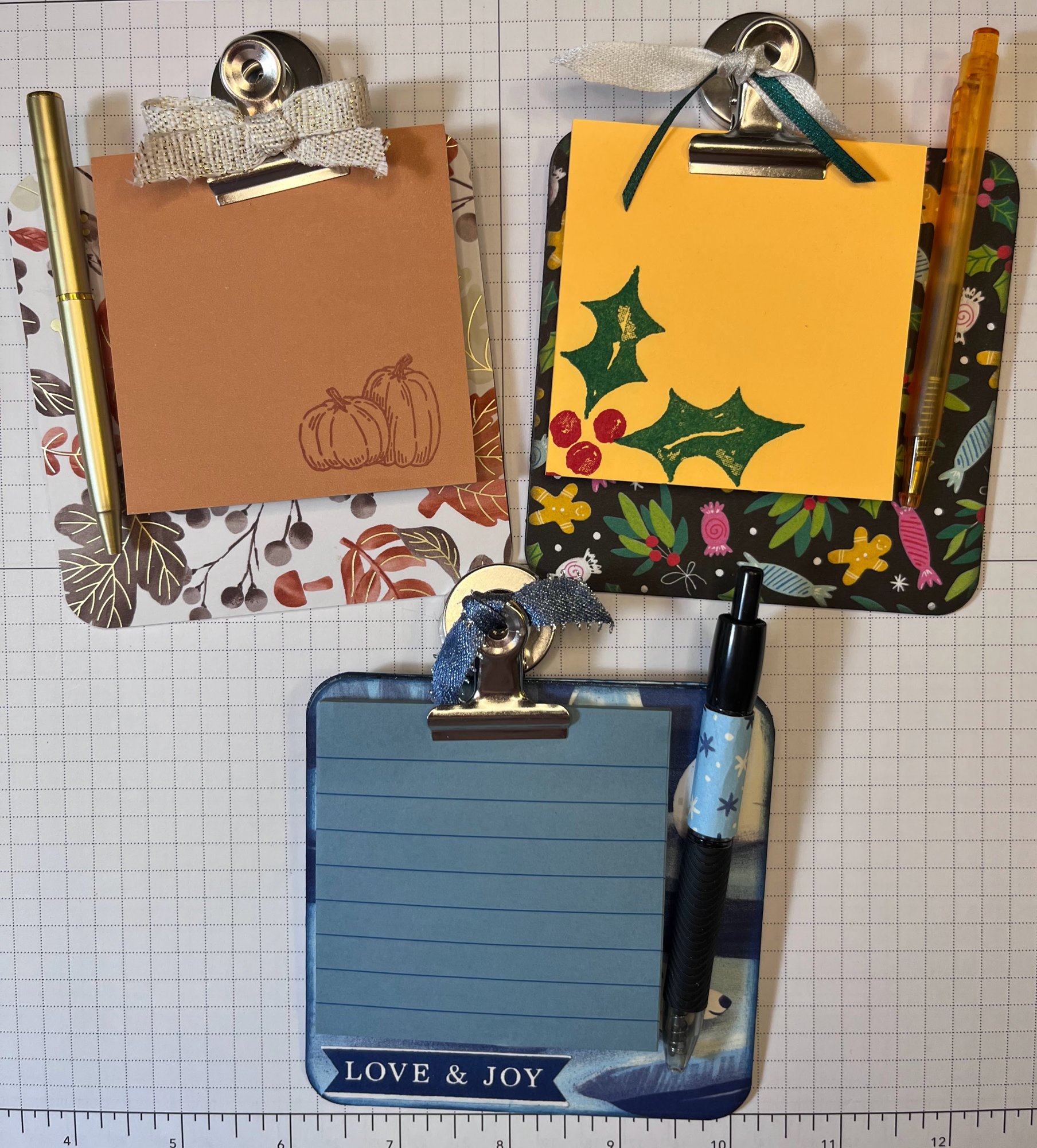 Three decorated mini clipboards with notepads and matching pens, in autumn, holly, and winter blue patterns